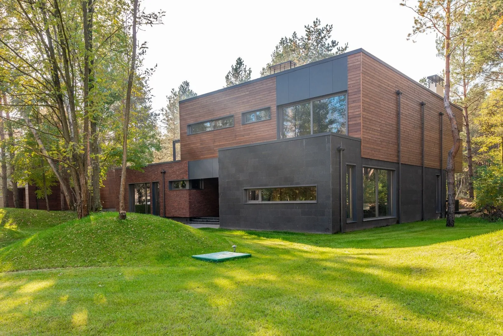 House of stacked blocks in larch boarding, brick and dark stone, set on a grass slope among tall pines.