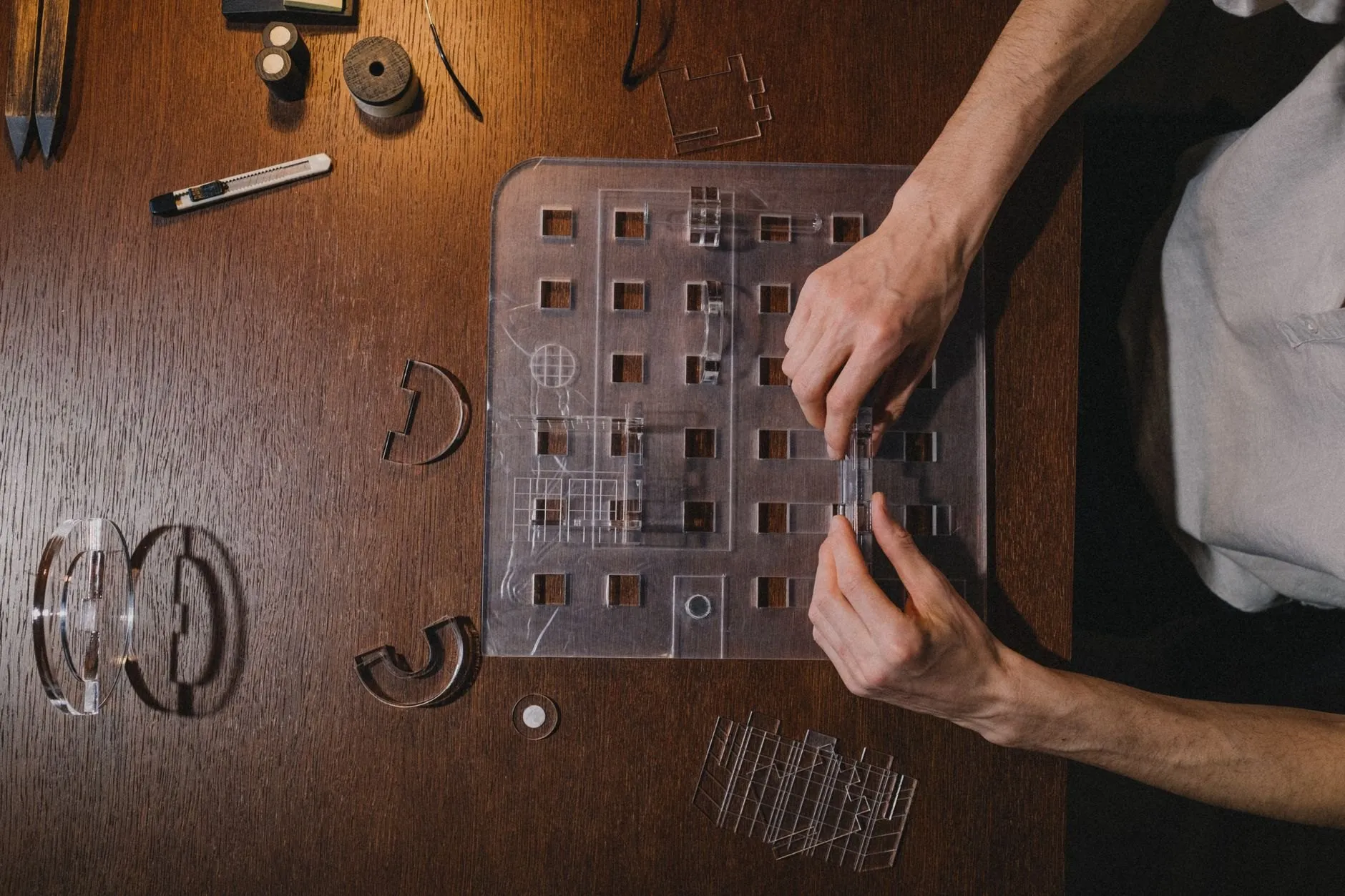 Hands assembling a clear acrylic model of a building facade on a dark wooden desk, cut pieces laid out around it.