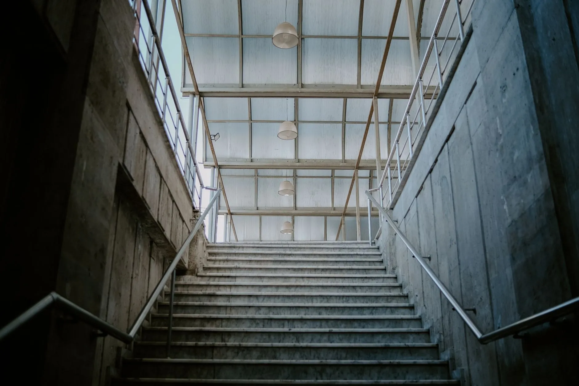 Stone stair rising between concrete walls towards a pitched glass roof with pendant lights.