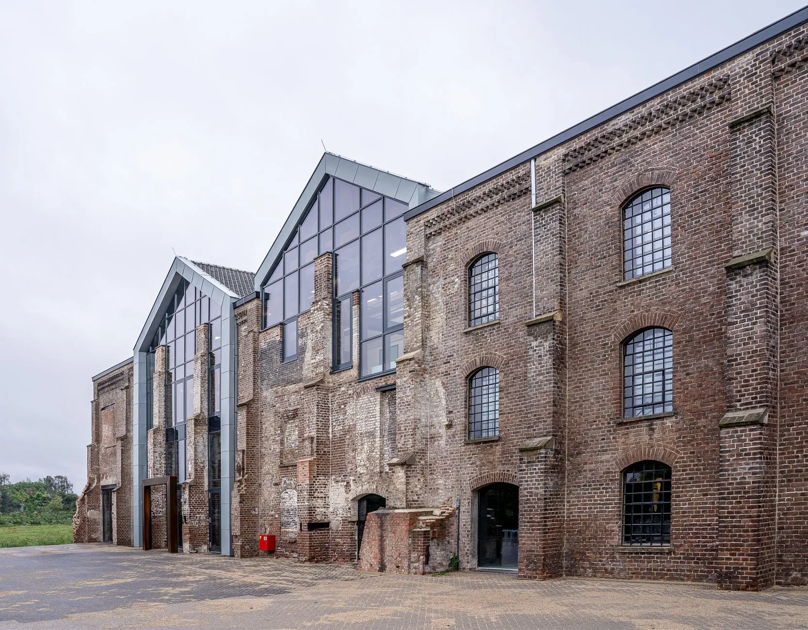 Weathered brick warehouse with two new glazed gables set into its end wall, under an overcast sky.