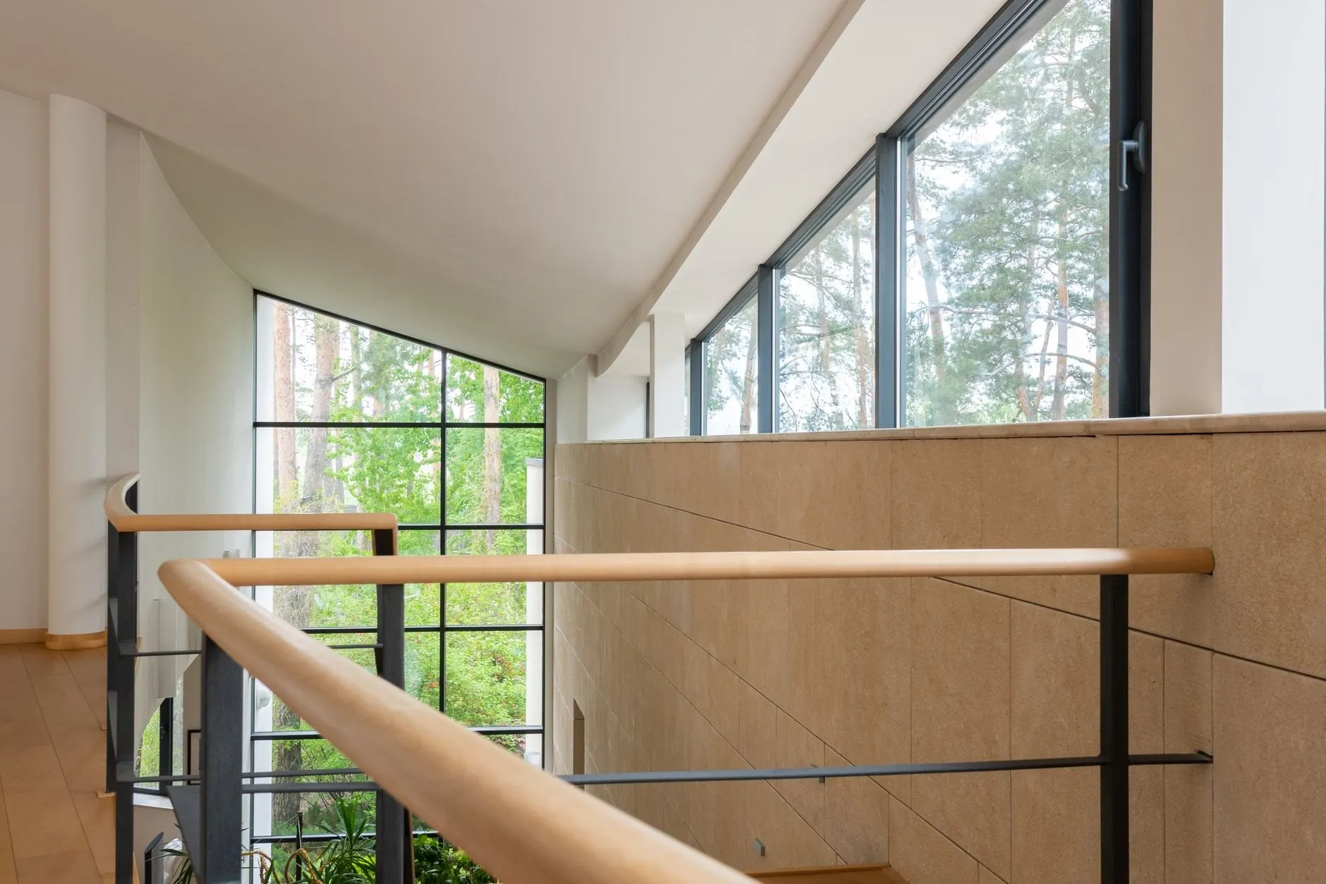 Split-level stair with an oak handrail and tall black-framed windows looking onto trees