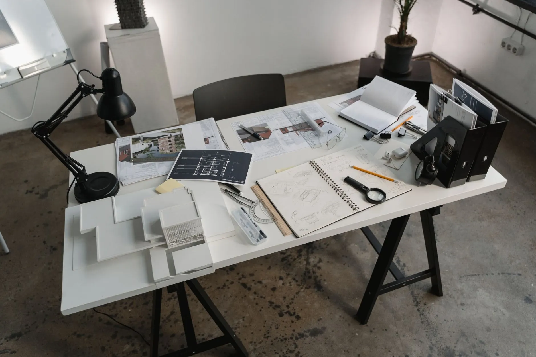 A studio desk with a black lamp, white card models, sketchbooks and printed plans on a concrete floor